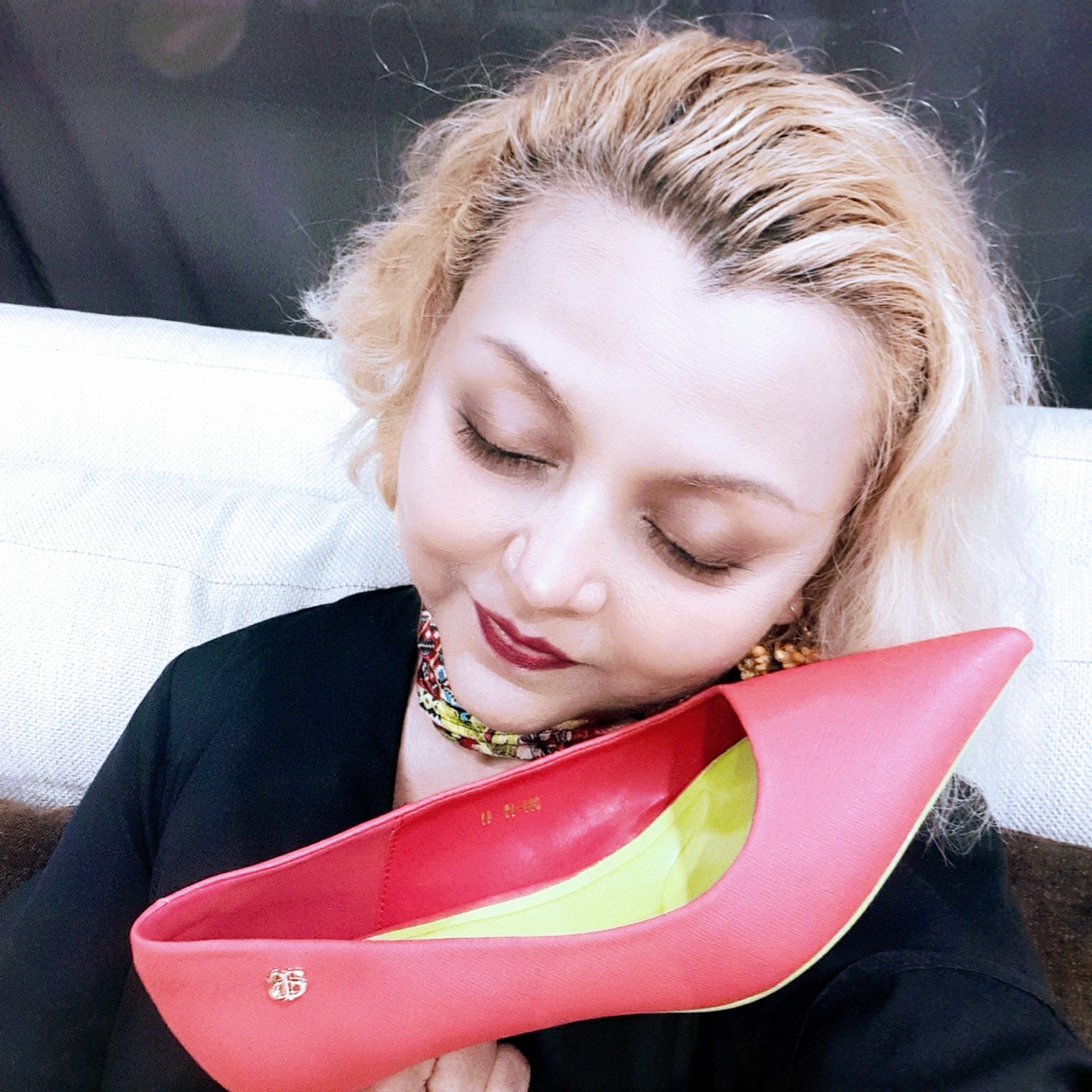 A woman with blonde, wavy hair and a colorful beaded necklace is seen holding a pink, pointed-toe, cruelty-free avarerarth pump with a green insole.